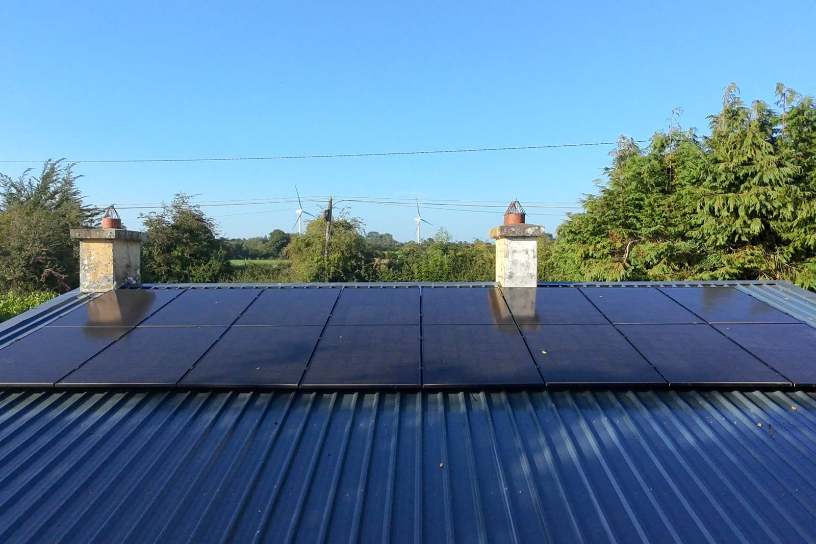 Solar panels across the corrugated roof of a farm building, with wind turbines on the horizon behind