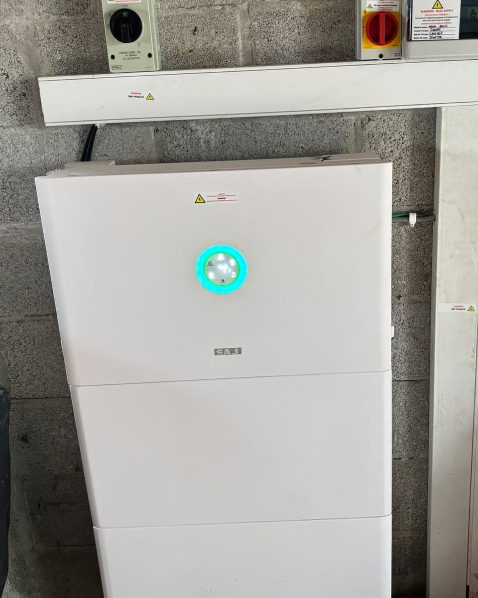 A hybrid inverter and stacked battery modules mounted on a garage wall, with the DC and AC isolators labelled on the shelf above