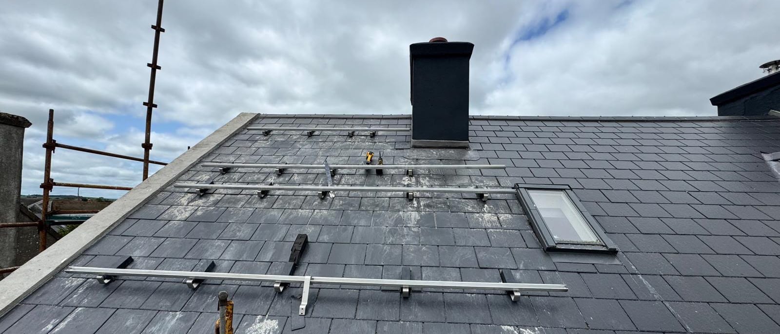 The slate roof mid-installation: mounting rails fixed to the rafters around the chimney and roof window, with no panels fitted yet